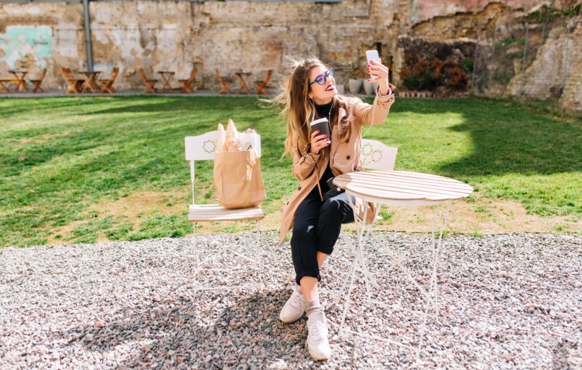 Attractive hipster girl wearing beige coat makes selfie for instagram profile during coffee break in